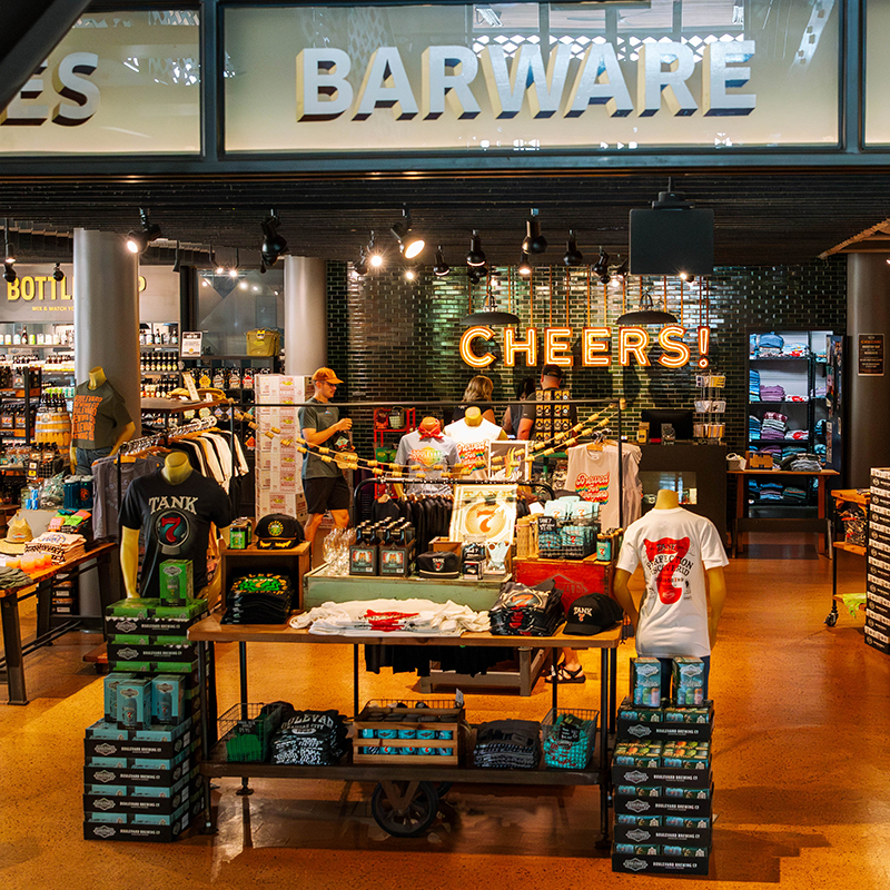 A lively store interior with a "CHEERS!" sign. Displays feature various products, including apparel with graphic designs and beer-related merchandise. Shoppers browse and staff assist near shelves stacked with items in a well-lit retail space.