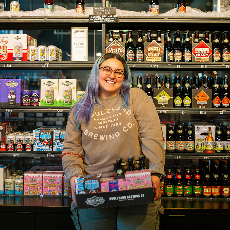 A person with long hair and glasses smiles while holding a caddy of Boulevard Brewing Co. beer bottles. They stand in front of a fridge stocked with various colorful beer and beverage labels.