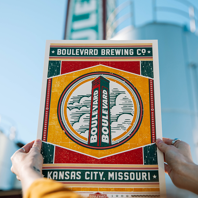 A person holding up a decorative Boulevard Brewing Co. poster with a vibrant design, featuring a brewery logo, vintage colors, and text reading "Kansas City, Missouri." The sky and part of a brewery building are visible in the background.