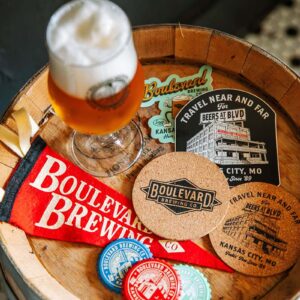 A glass of beer with foam on top sits on a wooden barrel, surrounded by various Boulevard Brewing Co. memorabilia including coasters, stickers, and a red pennant. The items feature logos, vintage designs, and branding of Boulevard Brewing Co., Kansas City, MO.