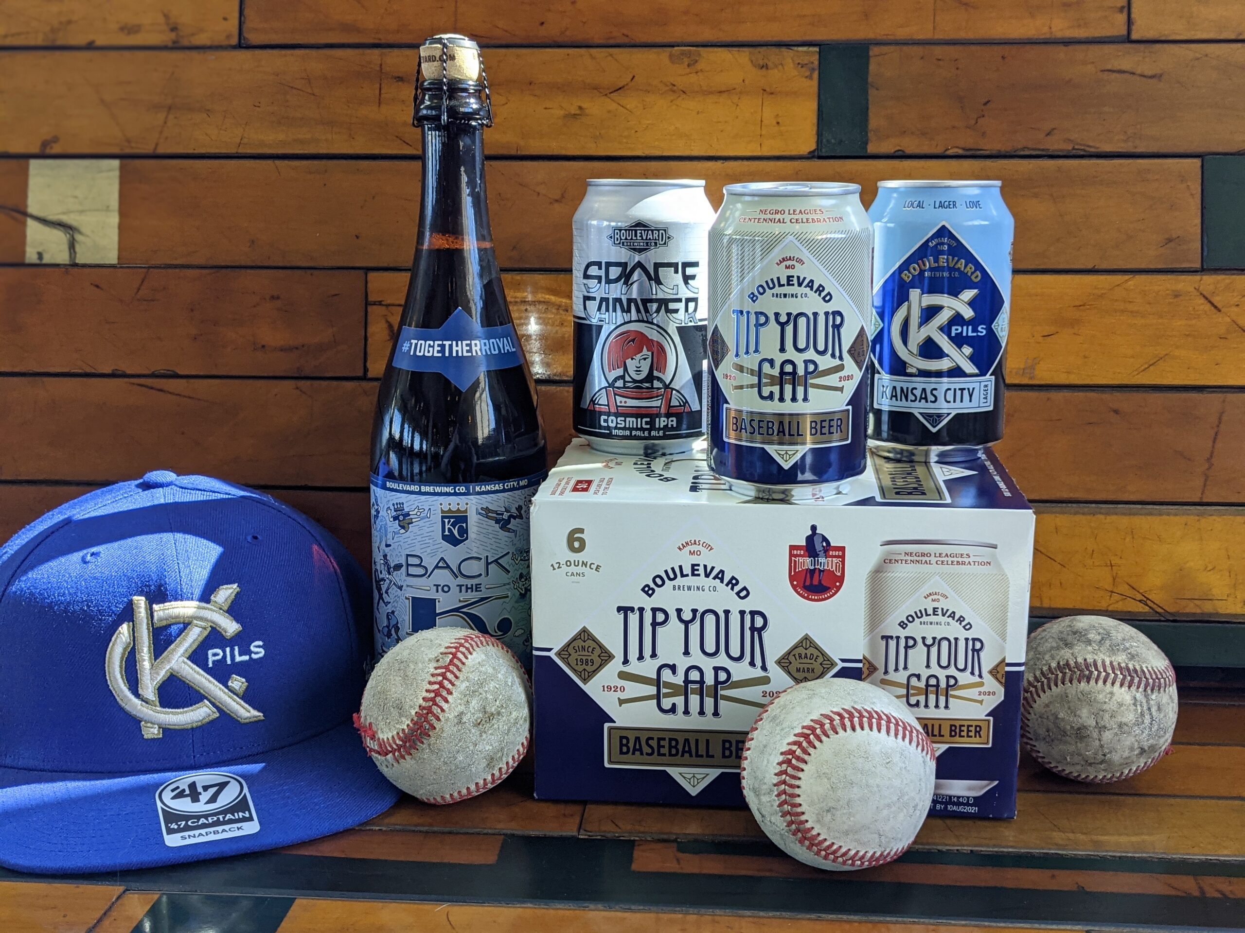 A collection of baseball-themed craft beer cans and bottles from Boulevard Brewing Company arranged on a wooden surface. A blue KC Pils cap and four baseballs surround the beverages, including "Tip Your Cap" and "Space Camper Cosmic IPA.