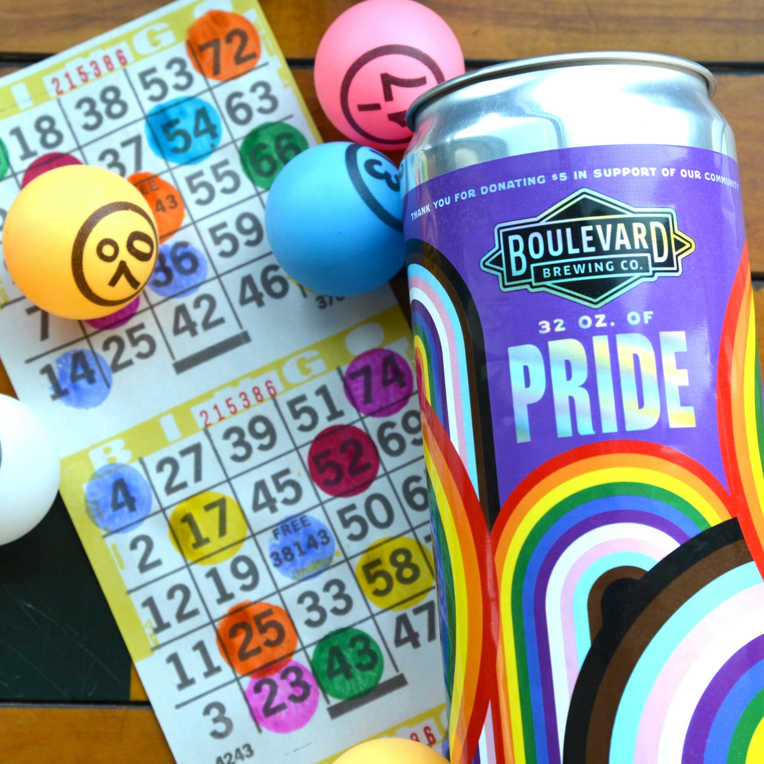 A can of Boulevard Brewing Co.'s Pride beer with colorful rainbow design is placed on a table. Nearby, there are bingo cards with various numbers marked and bingo balls in different colors, including one numbered 90, scattered around.