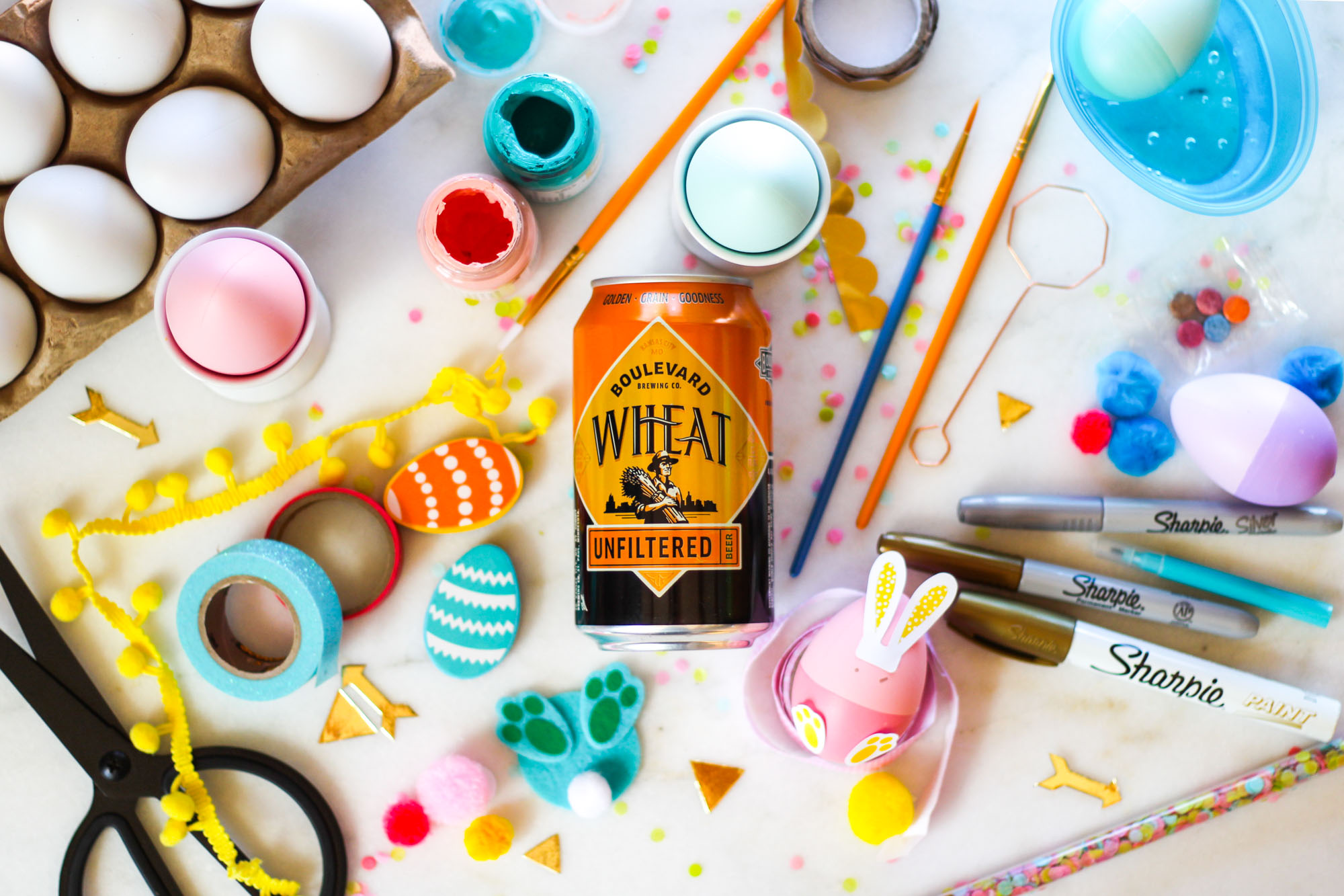 A colorful Easter craft setup with decorated eggs, paint, markers, scissors, washi tape, and pom-poms spread across a table. A can of Boulevard Wheat Beer is central among the art supplies and vibrant ornaments.