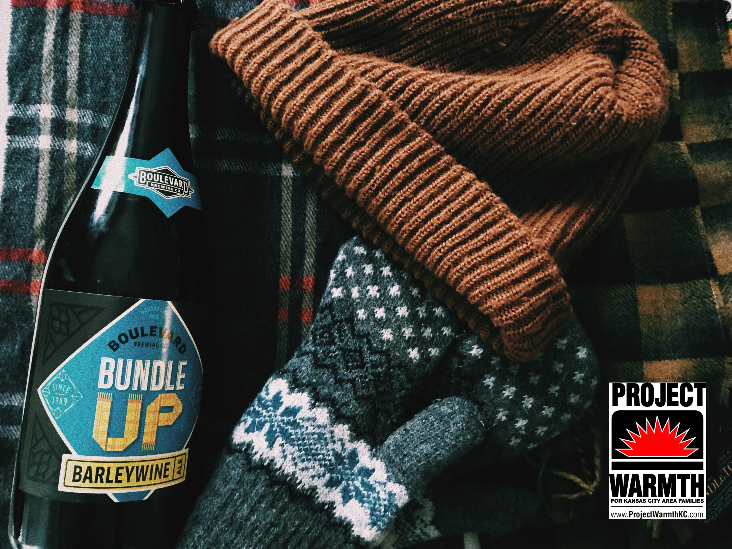 A bottle labeled "Bundle Up Barleywine" rests on a plaid fabric with a brown beanie and patterned gloves nearby. A logo for "Project Warmth" appears in the corner.