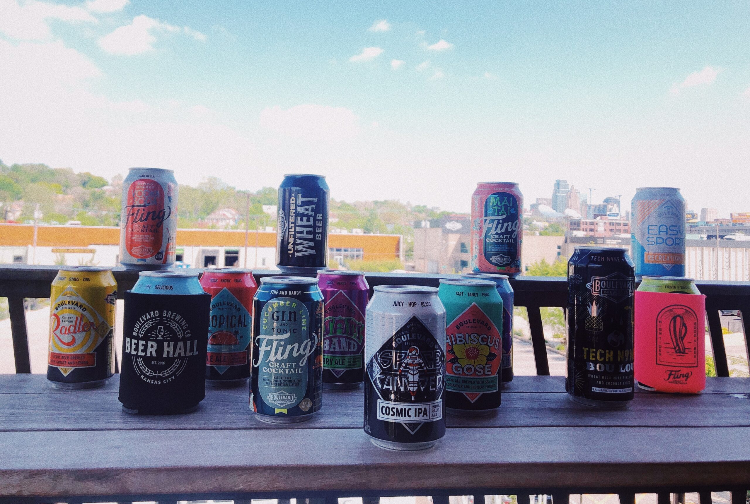 A variety of colorful beverage cans arranged on a wooden table with a cityscape and blue sky in the background.