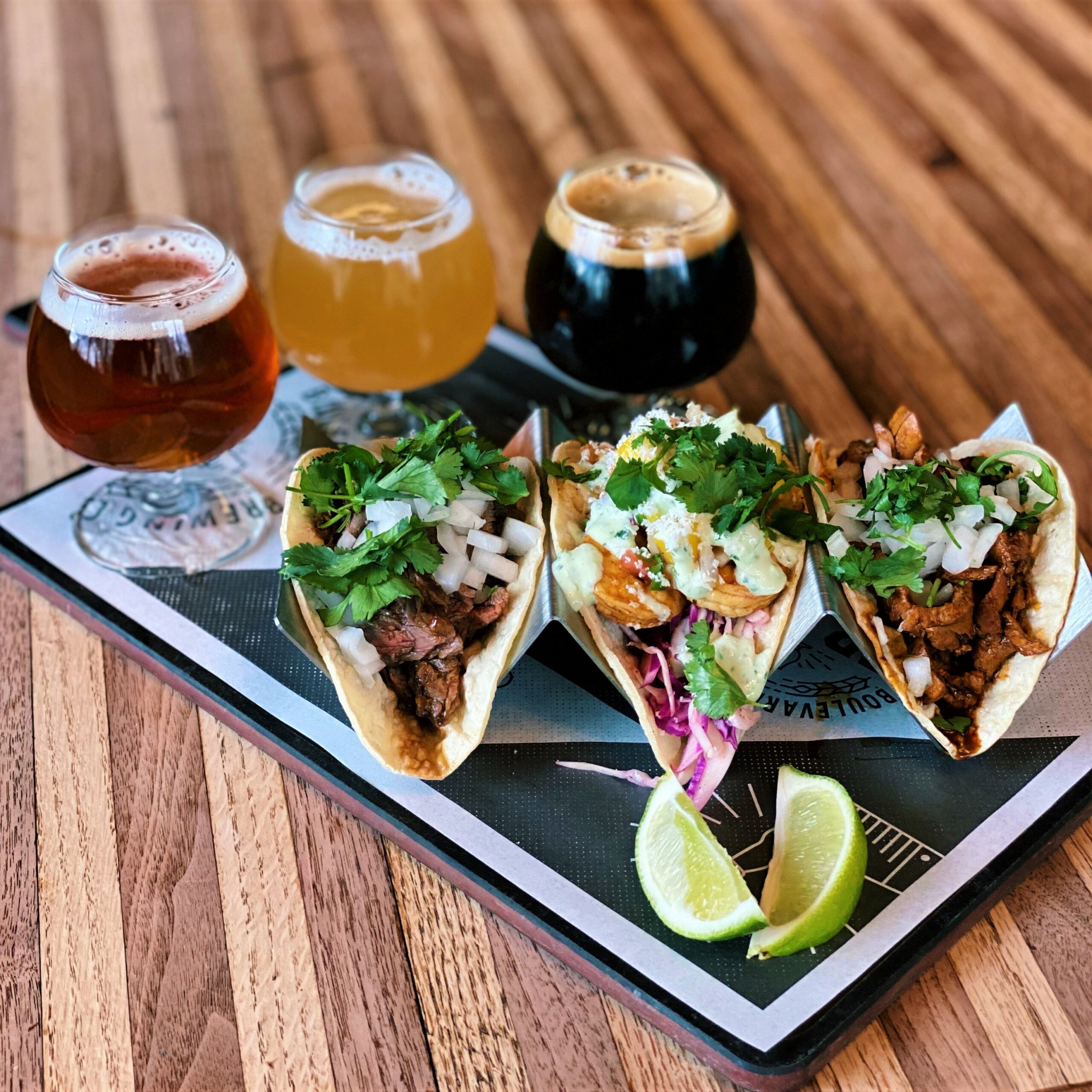 A tray with three tacos filled with meat, onions, and cilantro, accompanied by lime wedges. Behind the tacos are three glasses of different types of beer, ranging from light to dark. The scene is set on a wooden table.