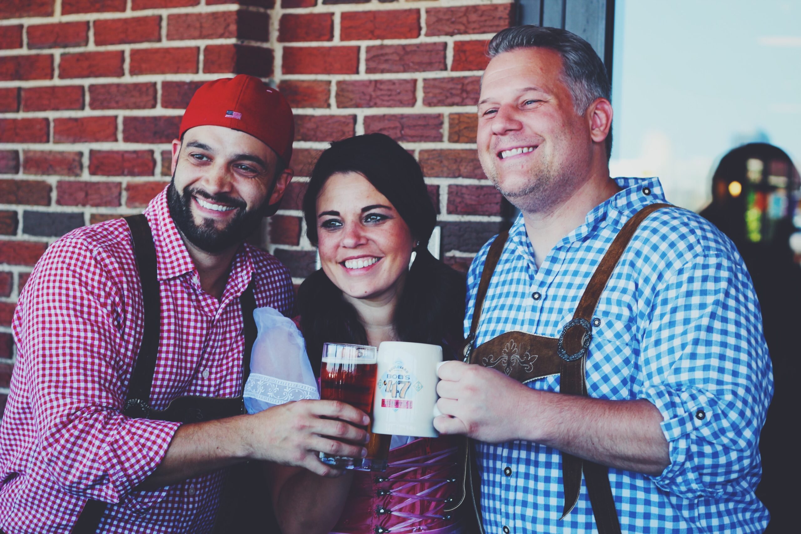 Three people in traditional Bavarian attire smile and hold drinks in front of a brick wall. The two men wear checkered shirts and lederhosen, and the woman, in a dirndl, stands between them.