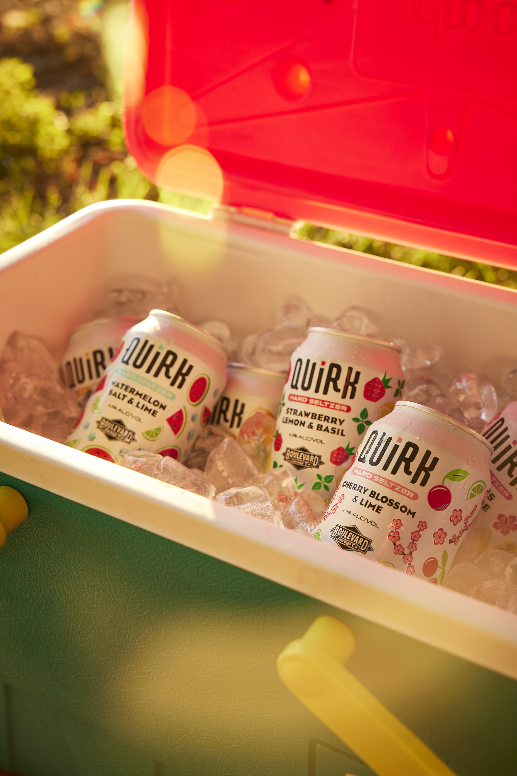 A cooler filled with ice holds cans of Quirk hard seltzer, with visible flavors including Watermelon Kiwi, Strawberry Lemon & Basil, and Cherry Blossom & Lime. The cooler has a green body, yellow handles, and an open red lid. Sunlight creates a warm glow.