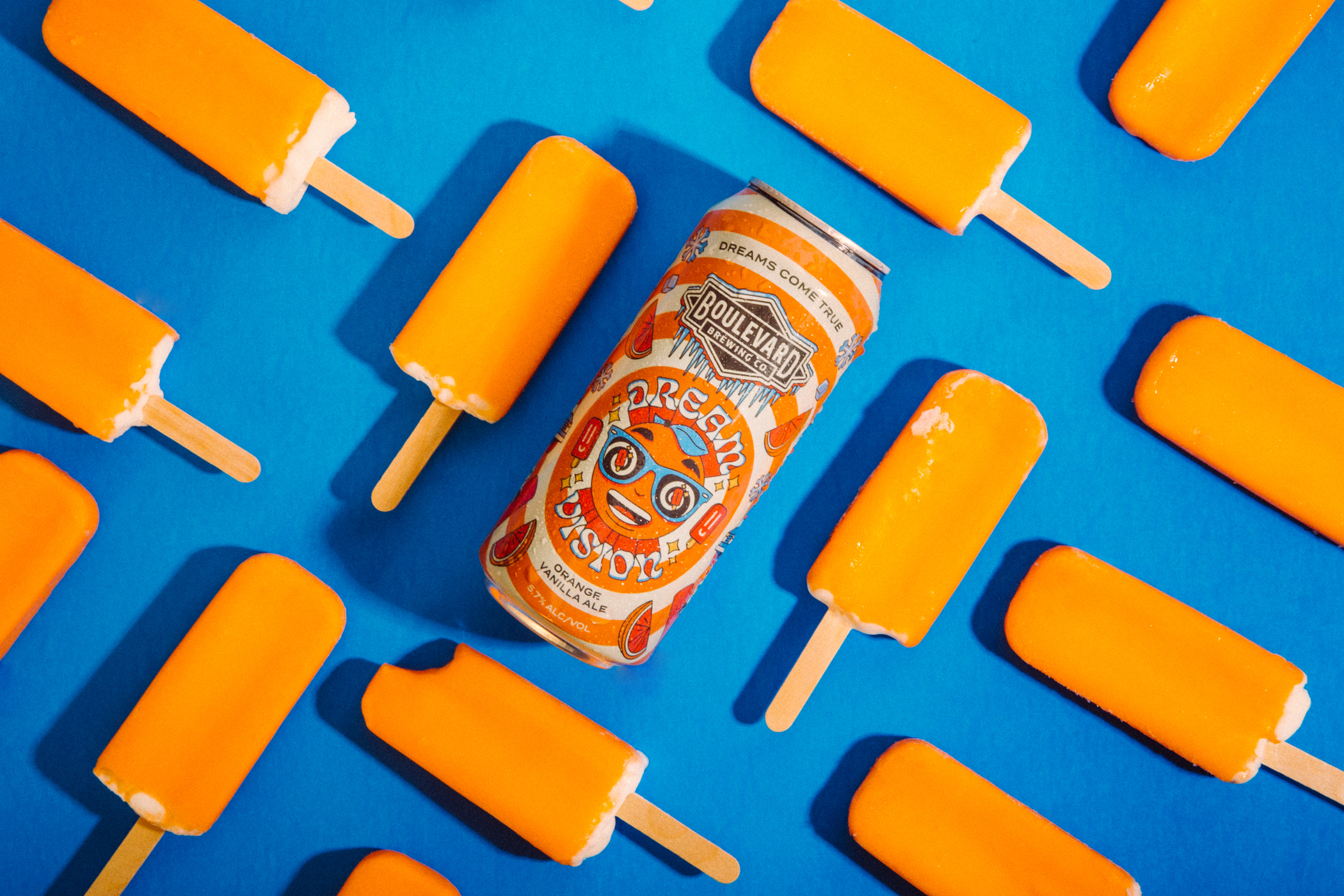 A can of Boulevard Brewing Co.'s Dream Vision beer is surrounded by vibrant orange popsicles on a bright blue background. The beer can features an artistic design with bold text.