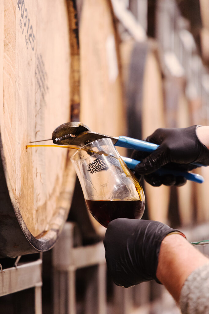 A person with black gloves uses a pipette to draw a dark liquid from a wooden barrel into a glass beaker with the logo "DOUBLE" on it. The background shows more barrels, suggesting a brewery or distillery setting.