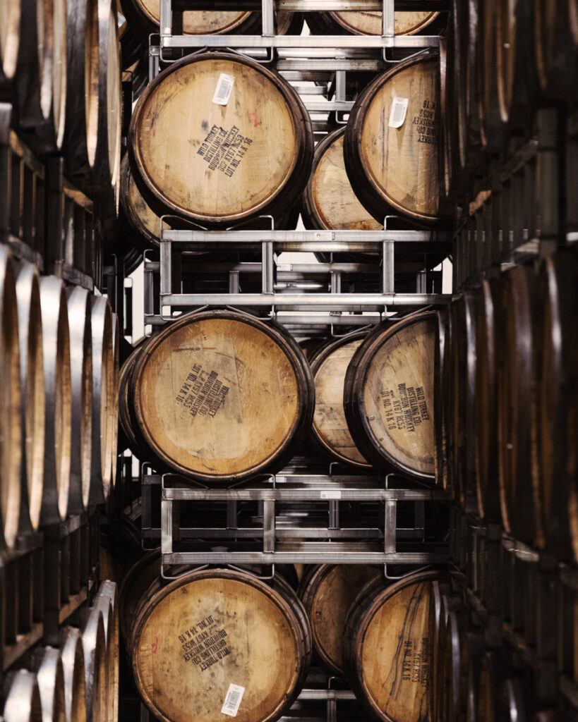 A narrow corridor between large wooden barrels stacked on metal racks, which are organized in rows from floor to ceiling. The barrels have labels on them and are slightly worn, suggesting use for aging beverages like wine or whiskey. The space is dimly lit.