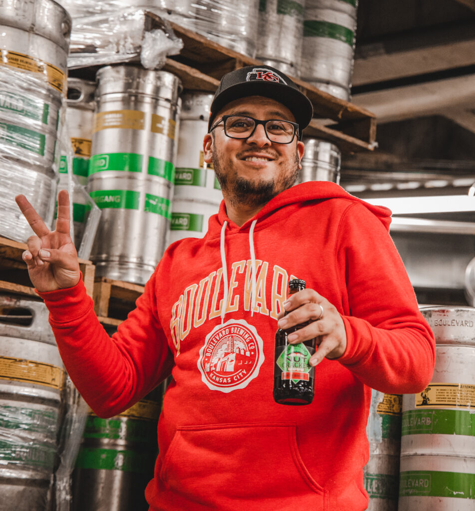 A person wearing a red hoodie with "Boulevard" on it and a black cap stands in front of stacked kegs, smiling and holding a beer bottle in one hand while making a peace sign with the other.