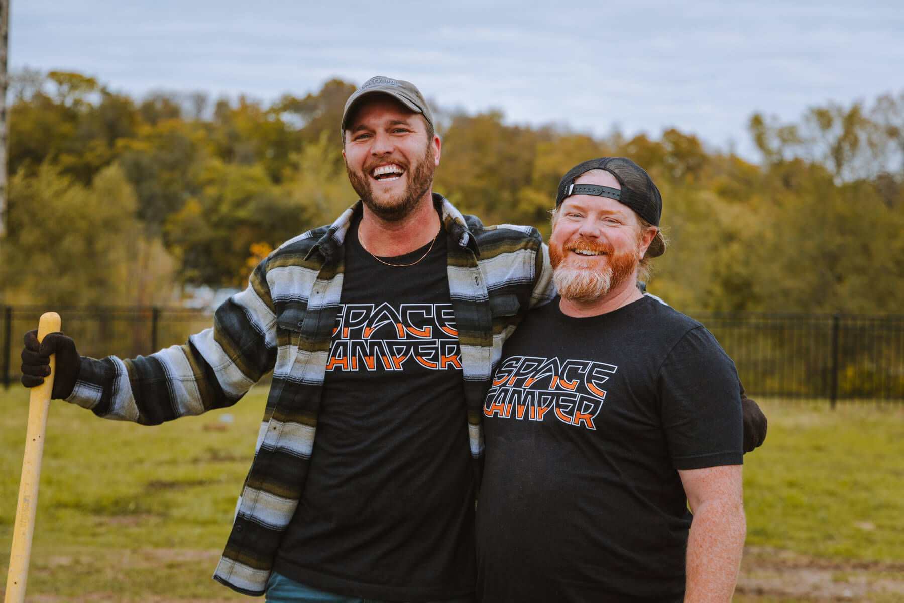 Two men wearing "Space Camper" T-shirts stand outdoors, smiling with arms around each other. One holds a shovel, and they are in a grassy area with trees and a fence in the background. They both are wearing caps; it's all about the relaxed and friendly atmosphere.