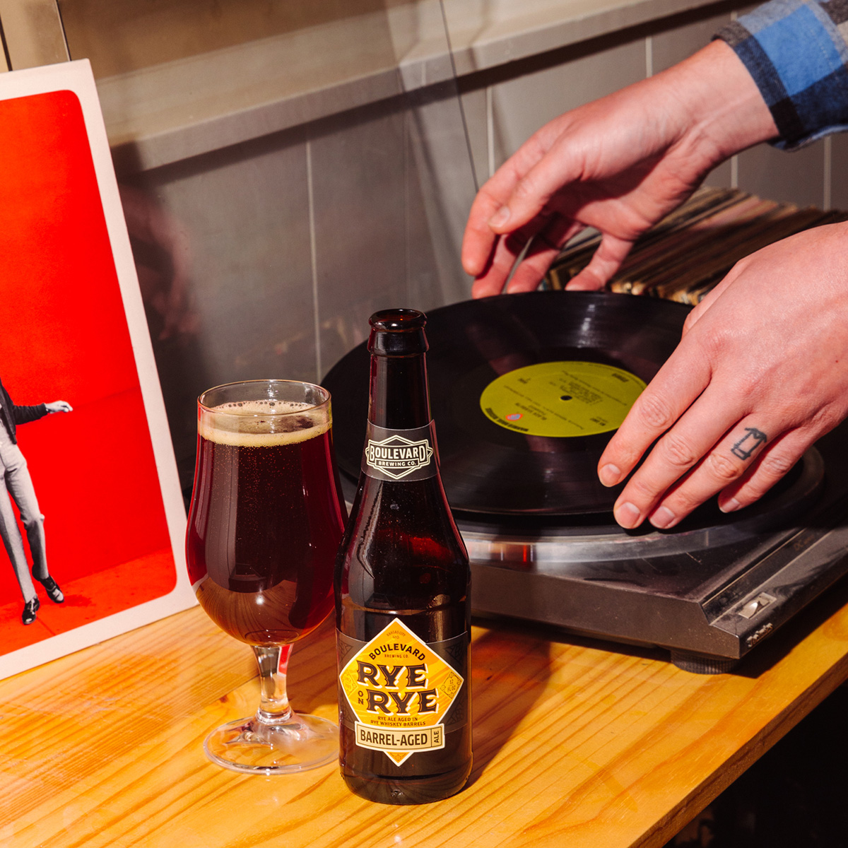 A person places a vinyl record onto a turntable. In the foreground, there's a bottle of Boulevard Rye on Rye beer and a filled glass next to it. The bottle label reads "Barrel-Aged." A red album cover with a figure in a red jacket is partially visible.