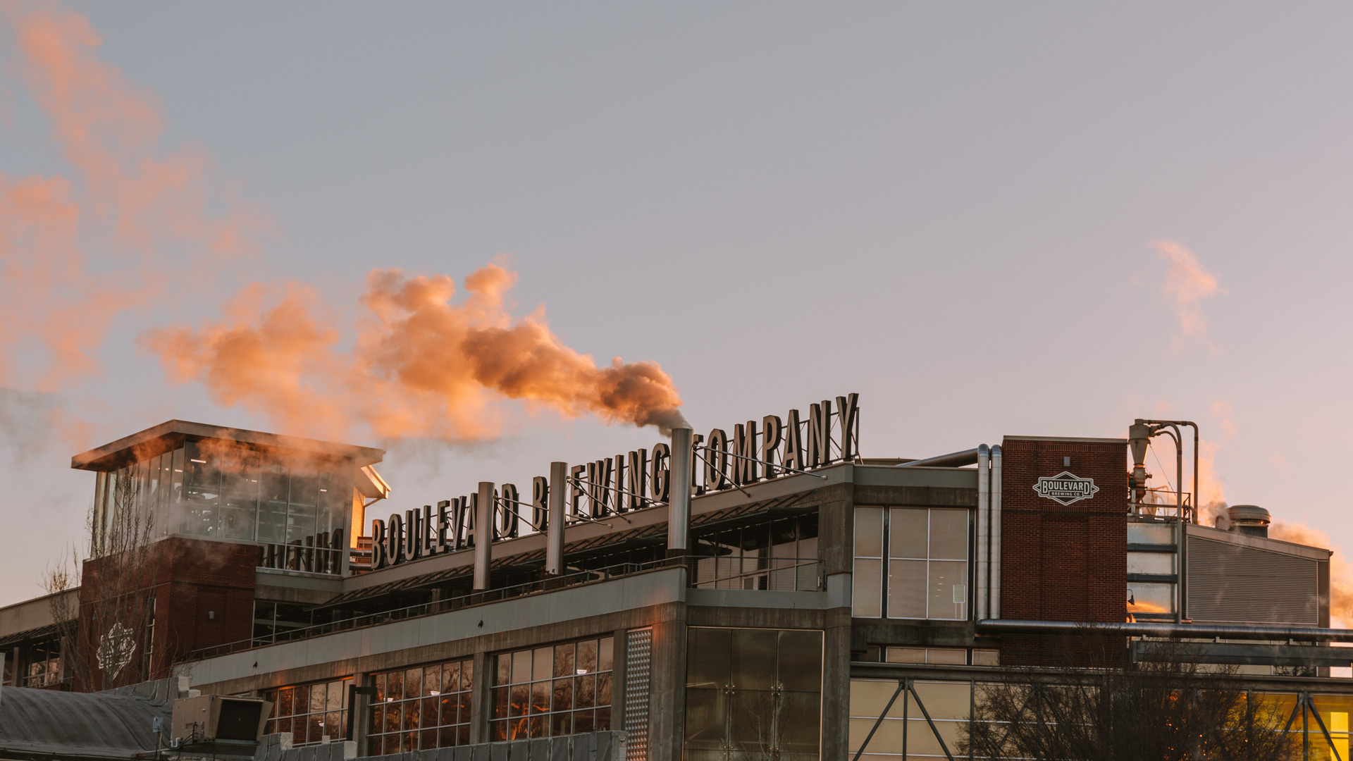 A brick brewery with a sign reading "Boulevard Brewing Company" emits steam at sunrise. The building, with its large windows and industrial design, tells a story about dedication and craftsmanship. The sky is clear with an orange hue, indicating early morning light.