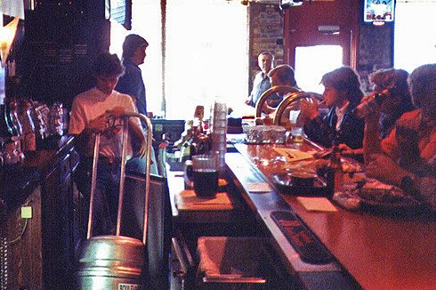 A bartender is seen behind the counter of a dimly lit bar, changing a keg. Several patrons are seated at the bar, drinking and conversing. The bar is adorned with various drink bottles, and a door is visible in the background, through which a few people are entering or exiting.