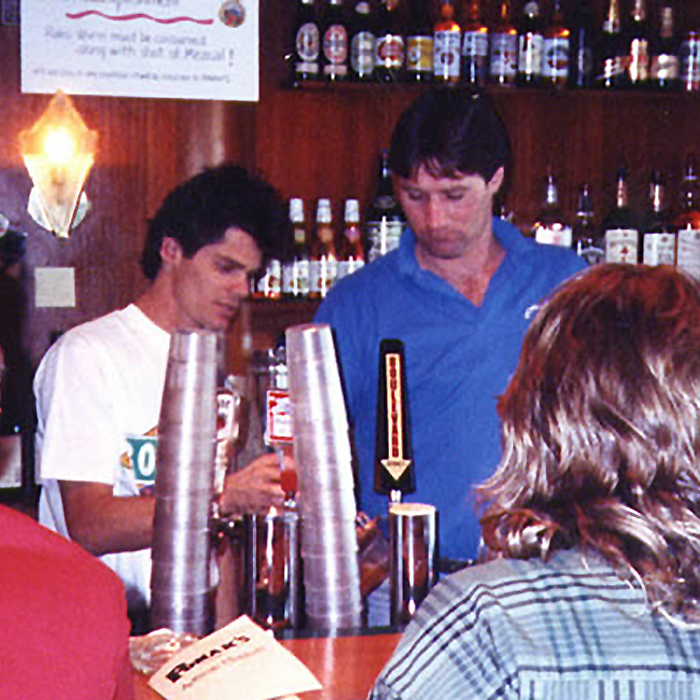 Two people standing behind a bar counter, organizing plastic cups in stacks. Various bottles of alcohol are displayed on the shelves in the background, and a customer with long hair in the foreground is facing the bartenders.