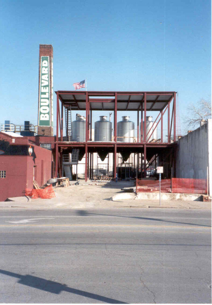 An industrial construction site featuring a metal framework of a building with several large tanks inside. A tall chimney with the word "Boulevard" written on it is on the left side. An American flag flies atop the structure, which is surrounded by fencing. This site is all about robust engineering and precision.