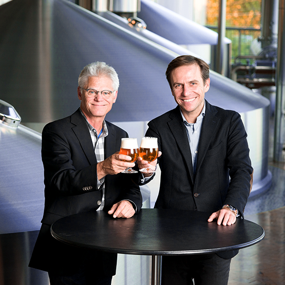 Two men in business attire, one with gray hair and glasses and the other with dark hair, smiling and holding glasses of beer. They are standing about a round black table with a brewery setup in the background.