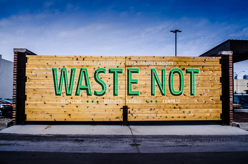 A large wooden sign mounted between two brick pillars with the words "Waste Not" prominently displayed in green letters. About it, smaller text reads, "Boulevard Brewing Co. Zero-Landfill Facility." Below, smaller text lists "Recycling," "Waste-to-Energy," and "Compost.