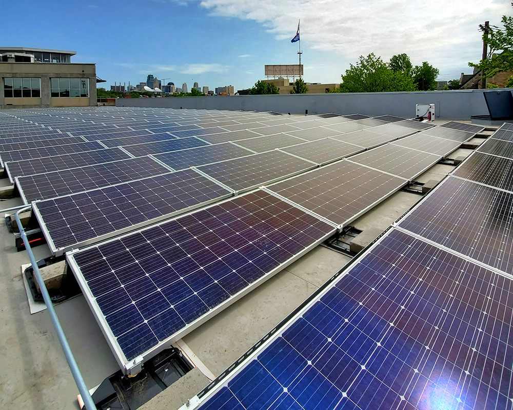 A rooftop covered with numerous solar panels angled towards the sky under a partly cloudy day. Buildings and trees are visible in the background, along with a blue flag near an elevated structure. The scene is about a renewable energy installation in an urban area.