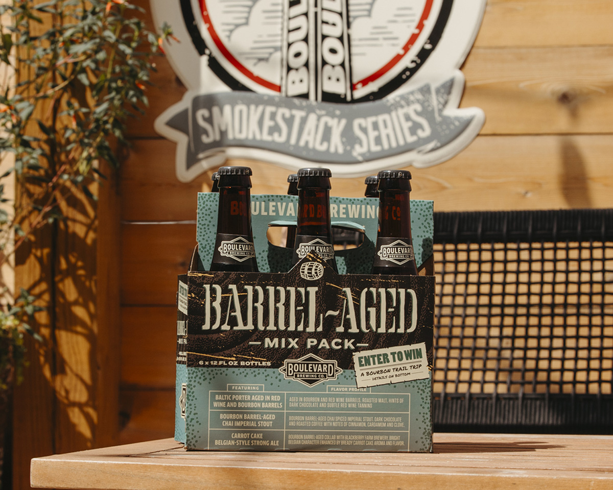 A six-pack of Boulevard Brewing Co.'s Barrel-Aged Mix Pack sits on a wooden surface. The packaging is green with black and gold accents, featuring three visible dark beer bottles. A sign with the brewery's logo and "Smokestack Series" is blurred in the background.