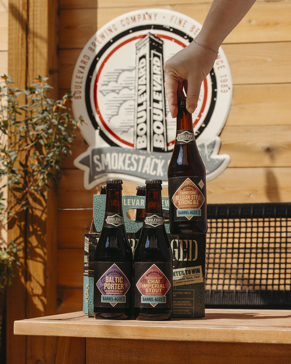 A hand holds a bottle of Belgian Style Strong Ale in front of a wooden wall with a Boulevard Brewing Company sign. The table has three Boulevard barrel-aged beers: Baltic Porter, Dark Sour Ale, and Belgian-style Strong Ale. A potted plant is on the left.