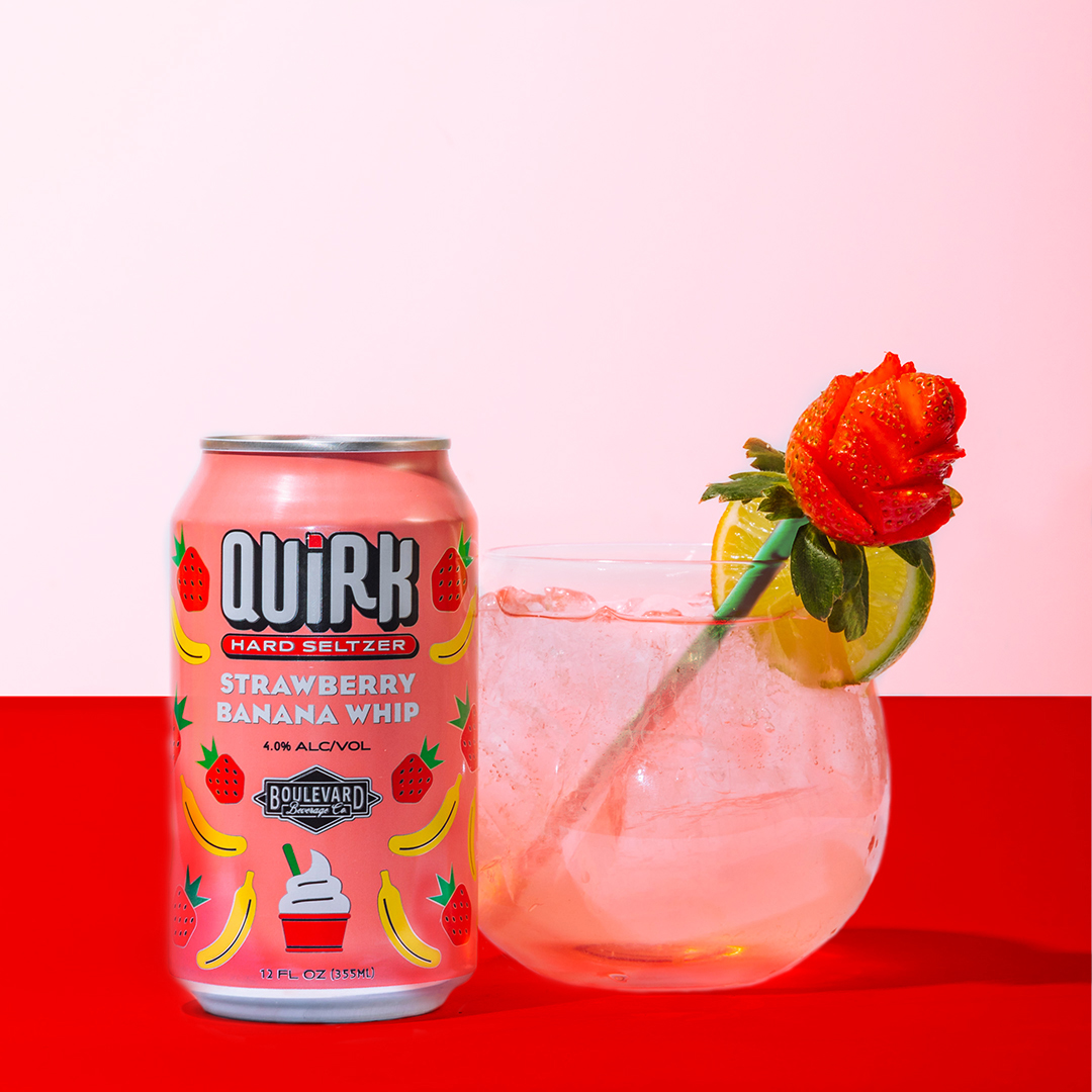 A can of Quirk Strawberry Banana Whip hard seltzer stands next to a glass of clear liquid with ice. The glass is garnished with a rose-shaped strawberry and a slice of lime on the rim. The background has a red bottom half and a white top half.