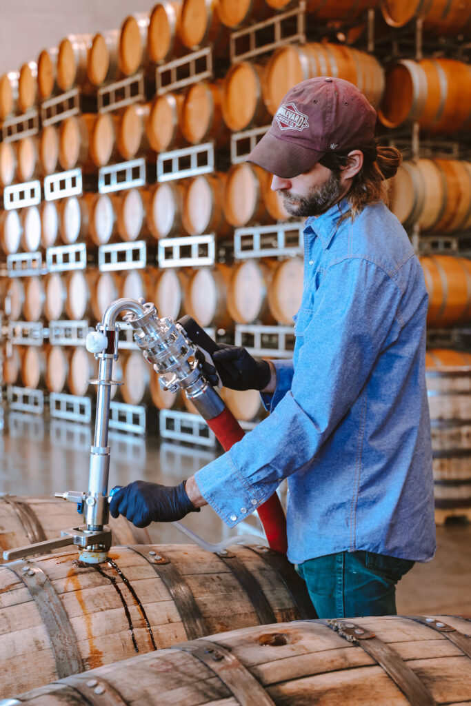 A person in a blue shirt and cap operates a hose connected to a wooden barrel in a warehouse filled with stacked barrels.