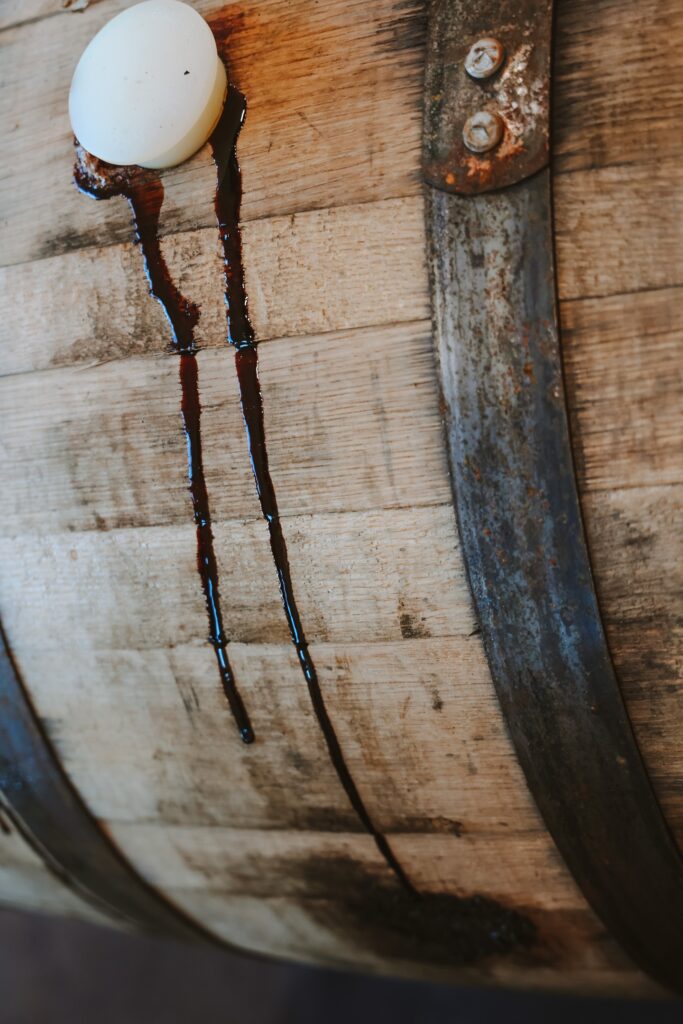 A close-up of a wooden barrel with metal bands. Dark liquid is dripping from an opening sealed with a white plug, running down the side of the barrel, creating elongated stains. The wood appears weathered and aged.