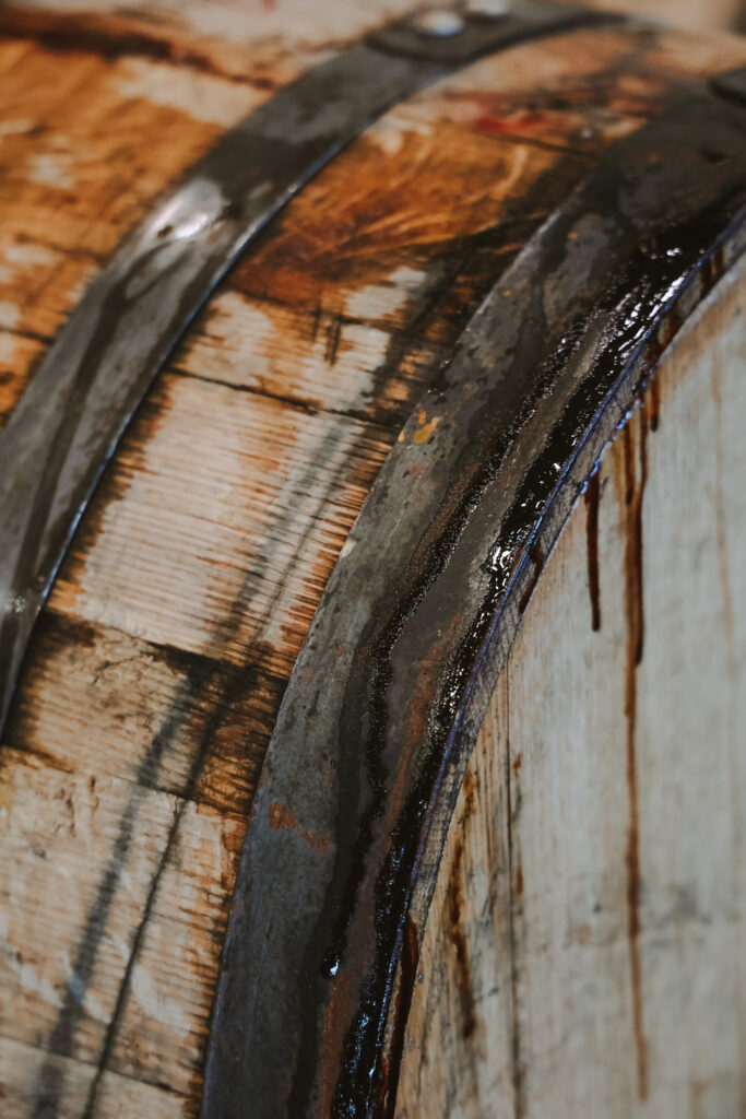 Close-up of an aged wooden barrel with dark metal bands, stained by a dark liquid seeping over the sides. The wood has a weathered appearance, highlighting the texture and grain. The image suggests the barrel might be used for aging beverages like wine or whiskey.