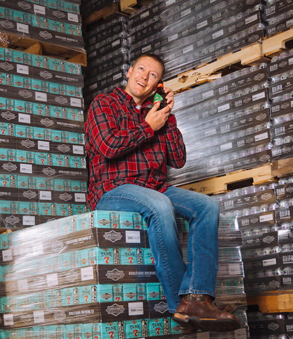 A person in a red plaid shirt and jeans sits on stacks of boxes, holding a bottle. Surrounding them are large pallets of similar boxes, suggesting a warehouse or storage setting. The person appears content and relaxed.