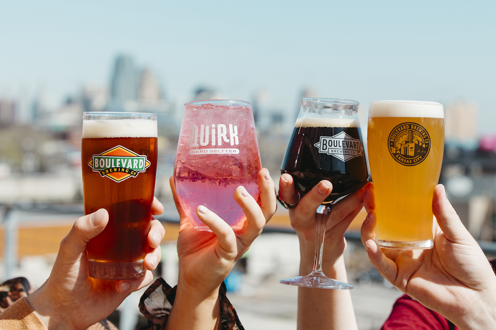 Four hands holding different types of beverages in branded glasses towards the camera, with a city skyline blurred in the background. The drinks include Pale Ale, Quirk hard seltzer, Bourbon Barrel Quad, and Unfiltered Wheat.