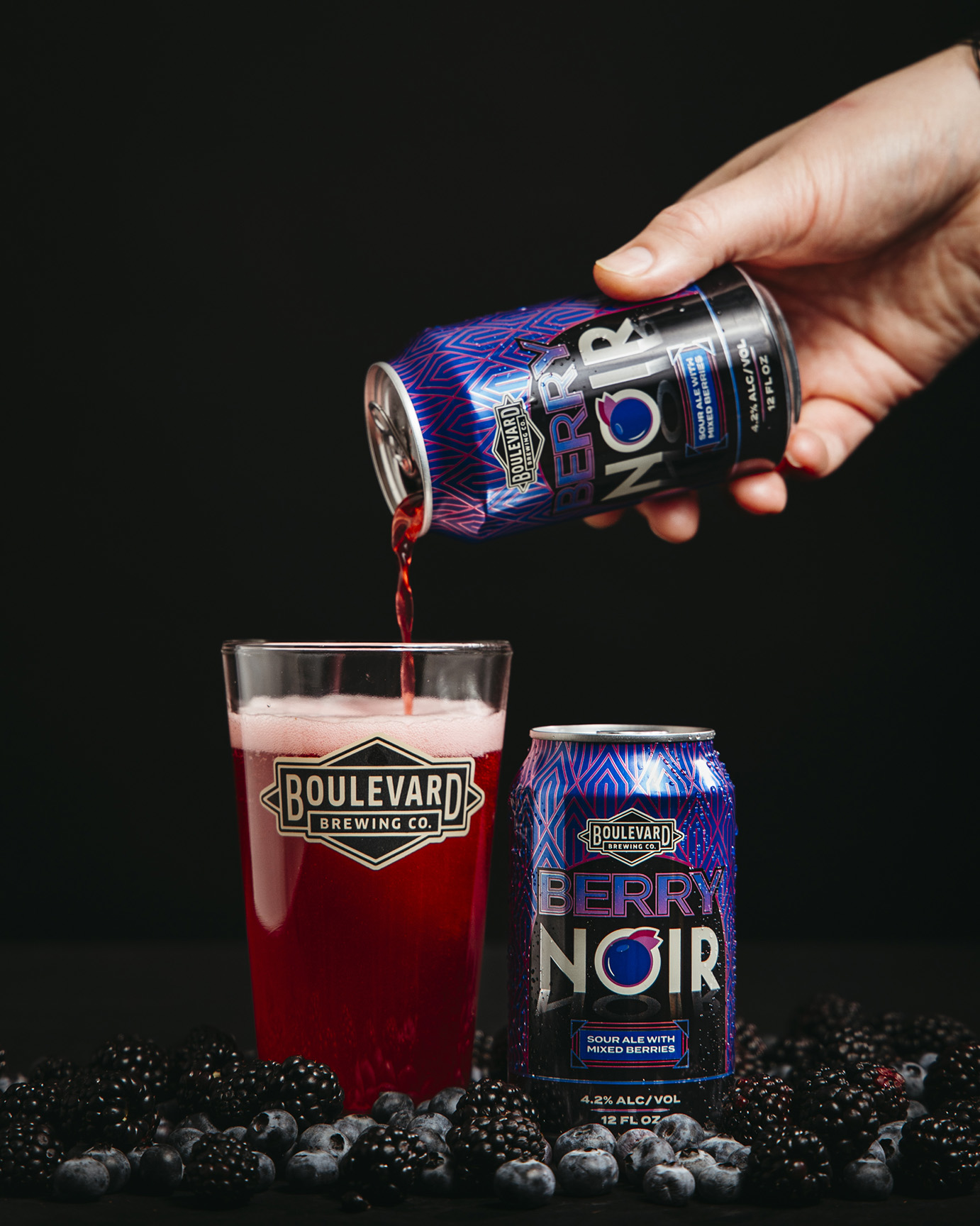 A hand pours Berry Noir beer from a Boulevard Brewing Co. can into a branded glass, surrounded by fresh blackberries and blueberries, against a black background.