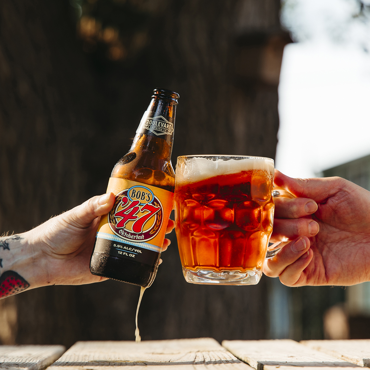 Two hands are shown, one holding a beer bottle labeled "Bob's 47" and the other holding a beer mug filled with amber beer. They are clinking together in a cheers gesture outdoors on a sunny day, with blurred trees in the background.