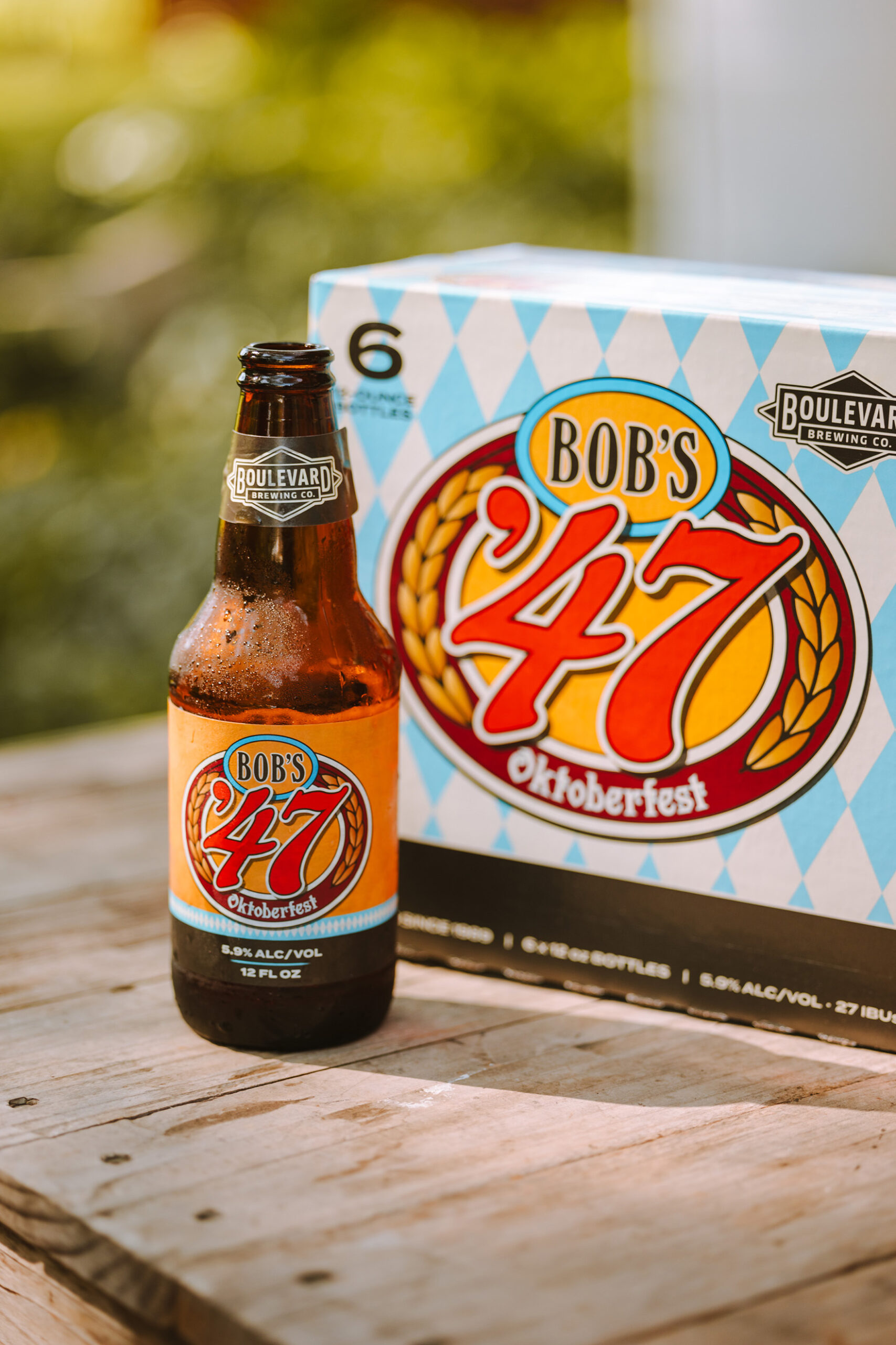 A bottle and a six-pack box of Bob's '47 Oktoberfest beer sit on a wooden surface. The label and box feature a festive design with red, yellow, and white colors. The background is blurred, showing a green outdoor setting.