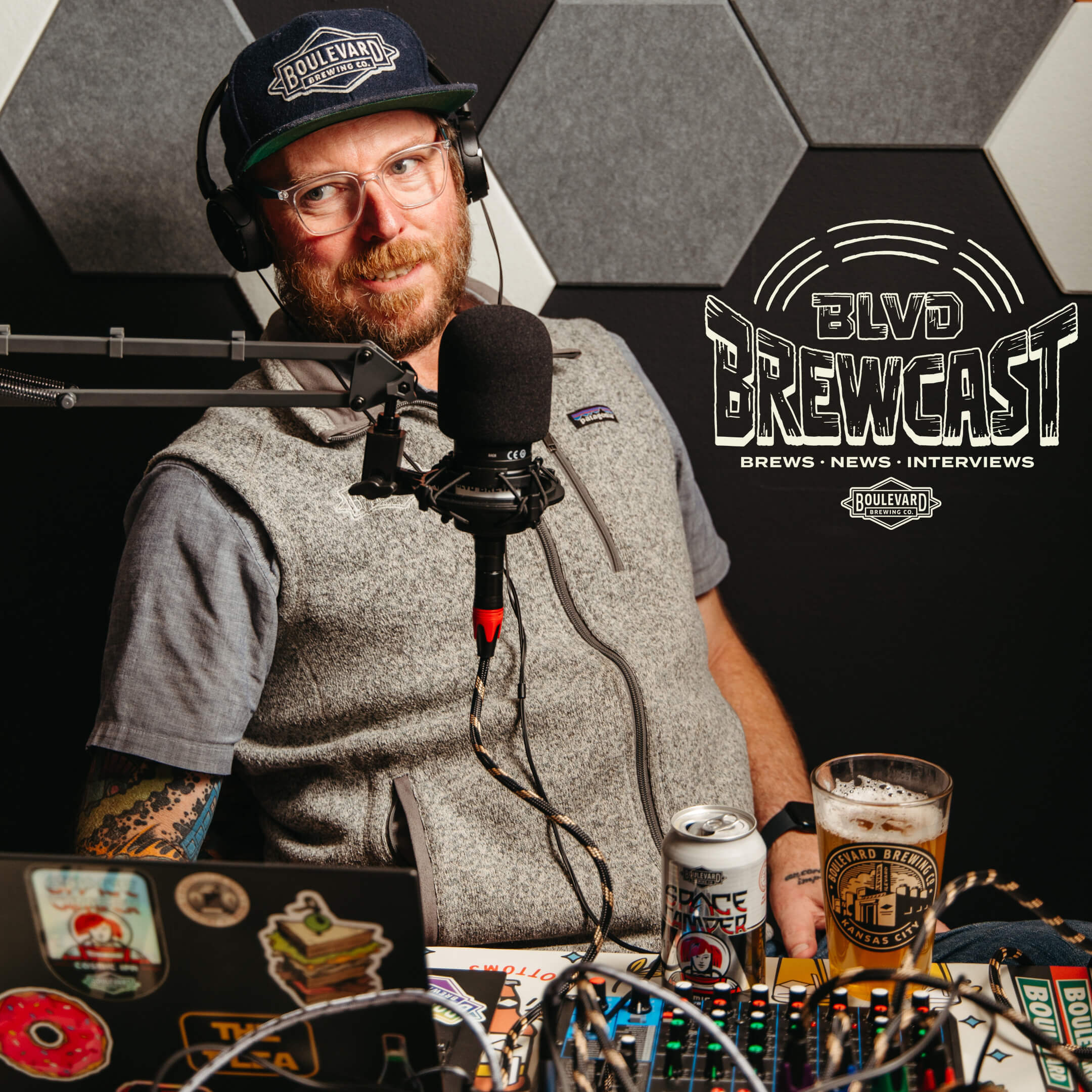 A bearded man wearing glasses and a baseball cap is sitting in front of a microphone in a recording studio. He is wearing headphones and speaking. A beer can, a beer glass, and a laptop with stickers are in front of him. The "BLVD Brewcast" logo is displayed.