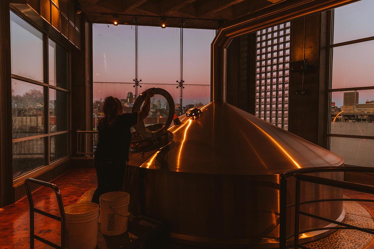 A person stands next to a large, shiny brewing tank in a dimly lit room with large windows. The sunset outside casts a warm glow over the scene. The individual appears to be inspecting or working on the tank, with various brewing equipment and buckets scattered about nearby.