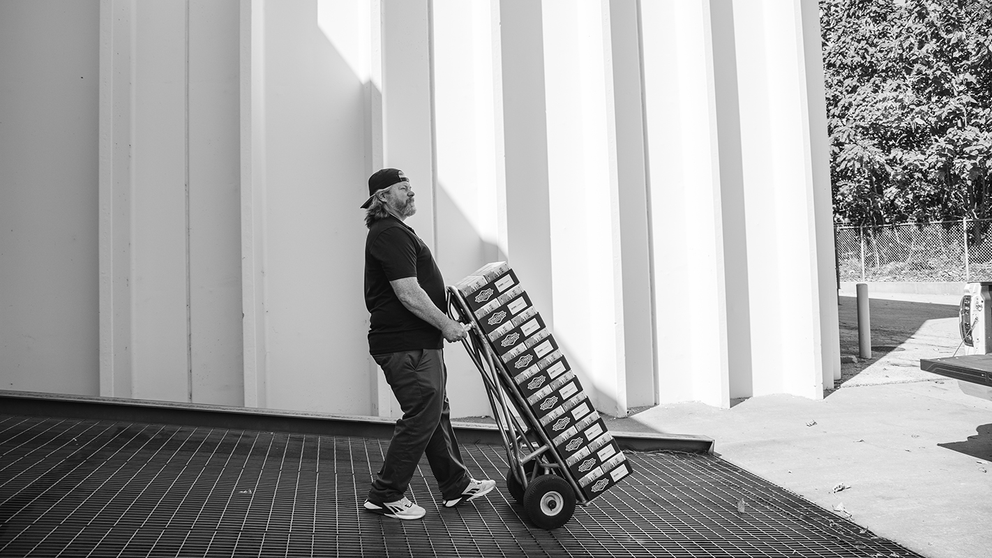 A person in casual clothes and a backward cap pushes a hand truck laden with boxes outside a large building. The black-and-white scene features sunlight casting strong shadows on the building's geometric facade. Trees are visible in the background, adding about a touch of nature to the urban setting.