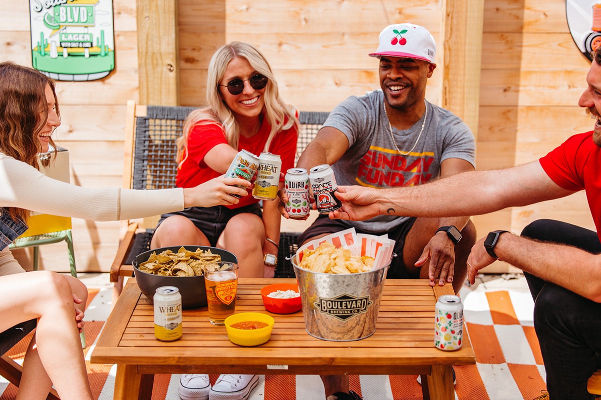 Four people, two women and two men, are sitting around a wooden table outdoors, smiling and clinking their cans of drinks together. About the table are a bucket of chips, a bowl of salsa, and a small bowl of cheese puffs. The setting appears casual and relaxed.