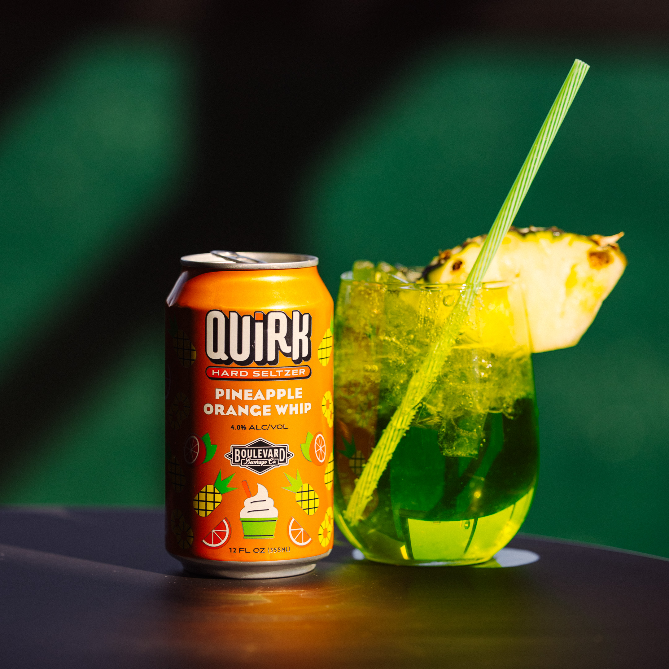 A can of Quirk Hard Seltzer, Pineapple Orange Whip flavor, sits next to a transparent cup filled with a greenish-yellow iced drink, garnished with a pineapple slice and a green straw. The can and glass rest on a dark surface against a green background.