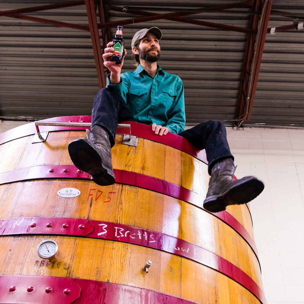 A man in a green shirt, black pants, and boots sits on top of a large wooden fermentation tank, smiling while holding up a bottle of beer. The background shows a metal roof and part of a white wall.