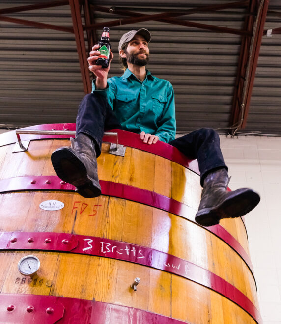 A man in a green shirt, black pants, and boots sits on top of a large wooden fermentation tank, smiling while holding up a bottle of beer. The background shows a metal roof and part of a white wall.