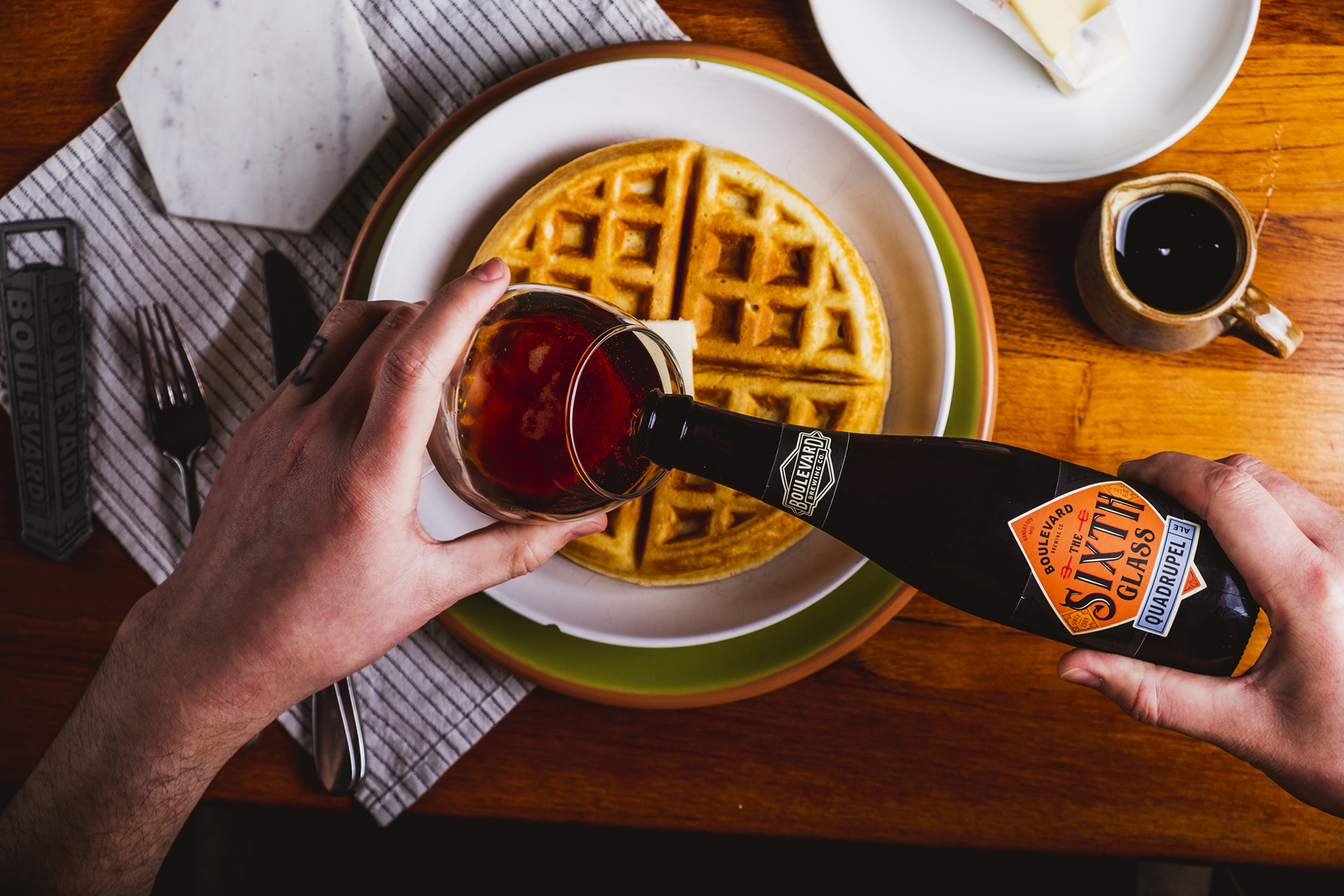 A person's hands are seen pouring a bottle of maple syrup over a stack of waffles placed on a white plate with a green rim. The table holds a small mug of coffee, a napkin with utensils, and a plate with butter slabs. The scene is set on a wooden table.