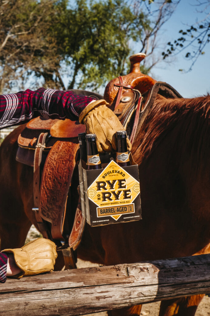 A person in gloves holds a four-pack of Boulevard Rye on Rye beer near the saddle of a brown horse. The background features trees and a clear blue sky. The focus is on the beer and the horse's body.