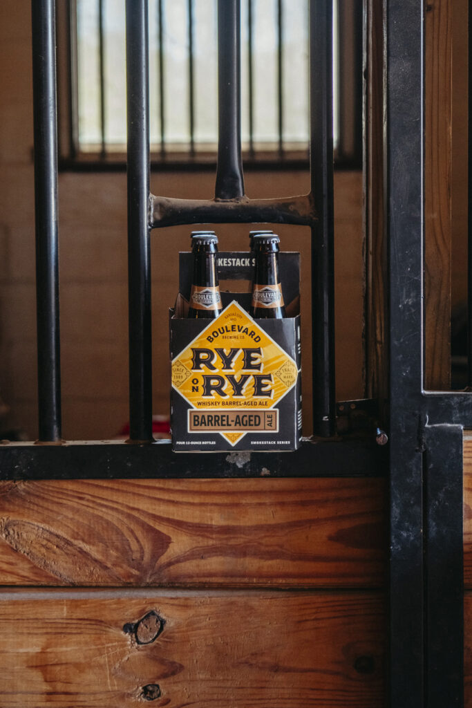 A pack of four Boulevard Brewing Co. Rye on Rye Barrel-Aged beer bottles is placed on a wooden ledge in front of vertical metal bars, likely part of a stable or rustic setting. The packaging is predominantly black and yellow, featuring the brand's logo and details.