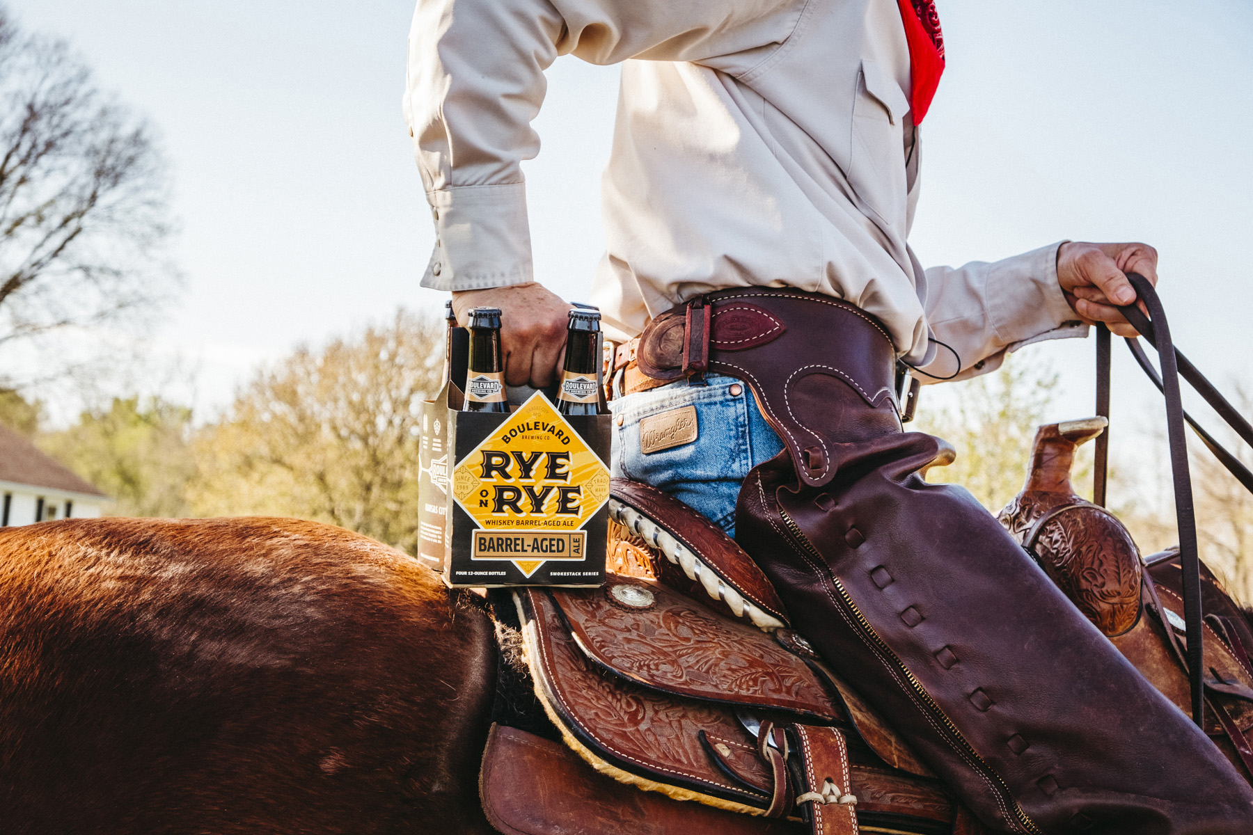 A person wearing a beige shirt and blue jeans, seated on a horse, holds a six-pack of Rye beer in their right hand. The rider is equipped with traditional cowboy gear, including chaps and a belt, with trees and bright sky in the background.