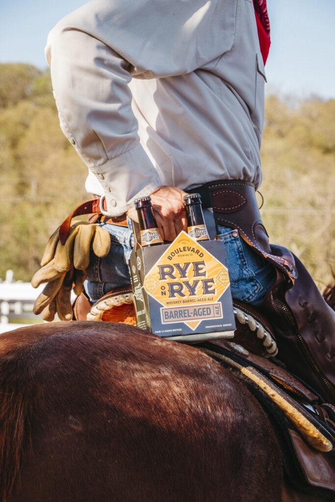 A person wearing jeans and a light-colored shirt sits on a horse, holding a six-pack of Boulevard Rye on Rye Barrel-Aged beer. The day appears sunny with trees in the background. The rider is also seen wearing gloves and a bandana.