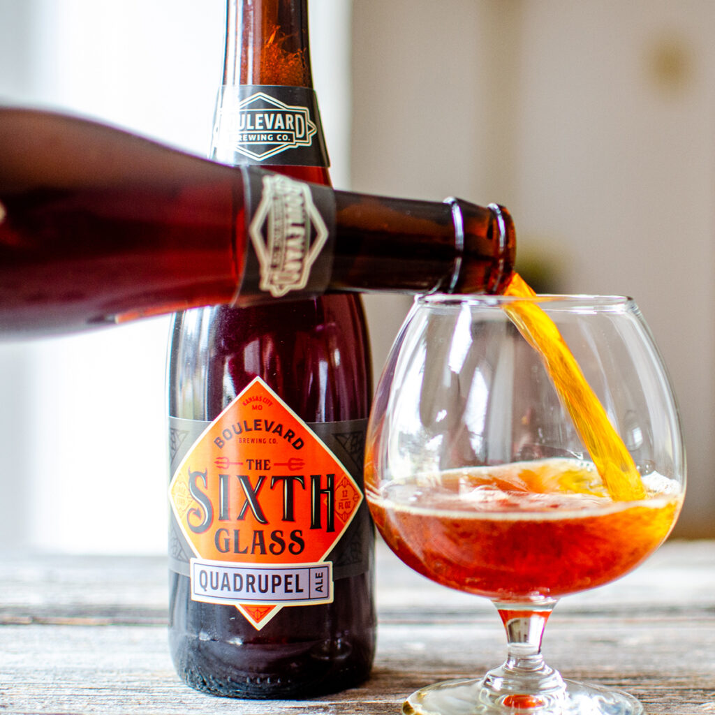 A glass of golden amber beer being poured from a Boulevard Brewing Co. bottle of The Sixth Glass Quadrupel Ale. The bottle is partially visible, showcasing its label, while the glass is nearly full with foamy beer. The setting appears to be indoors.