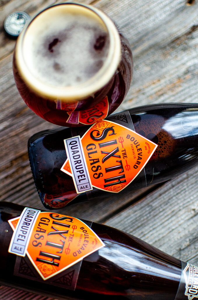 Top view of a wooden table with two bottles of Boulevard's The Sixth Glass Quadrupel Ale lying on their sides, and a filled glass of the ale with frothy foam on top. The labels on the bottles are prominently visible, featuring geometric designs.